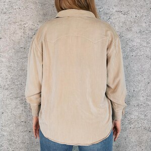 May include: A tan corduroy shirt with long sleeves, a button-down front, and a classic collar. The shirt features a Western-style yoke detail on the back and cuffs with button closures. The shirt is paired with blue jeans.