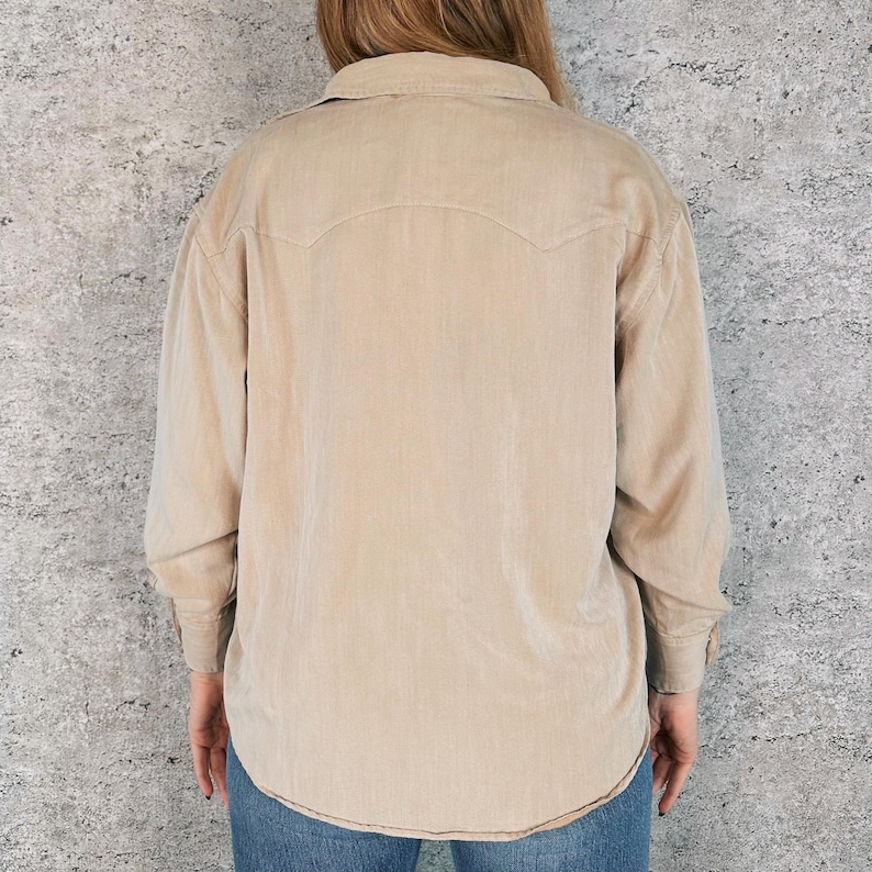 May include: A tan corduroy shirt with long sleeves, a button-down front, and a classic collar. The shirt features a Western-style yoke detail on the back and cuffs with button closures. The shirt is paired with blue jeans.