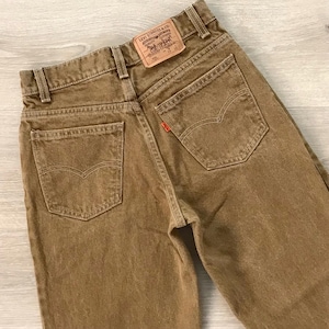 May include: Close-up of brown Levi Strauss & Co. jeans. The jeans have a classic five-pocket design with a small red tag on the back pocket. The Levi Strauss & Co. label is visible on the back waistband.