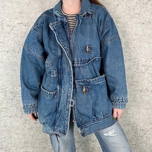 May include: A medium-wash blue denim jacket featuring a high collar, long sleeves with ribbed cuffs, and a front zipper closure. The jacket includes two large patch pockets on the front, each secured with a small wooden toggle button detail.