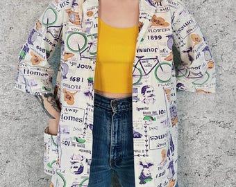 May include: Cream-colored blazer with a bicycle and newspaper print in green, purple, and orange. The blazer has a collared neckline, three-quarter sleeves, and two front pockets. The text on the blazer includes "FLOWERS 1899" and "Typewriter Bicycle Bell 10".