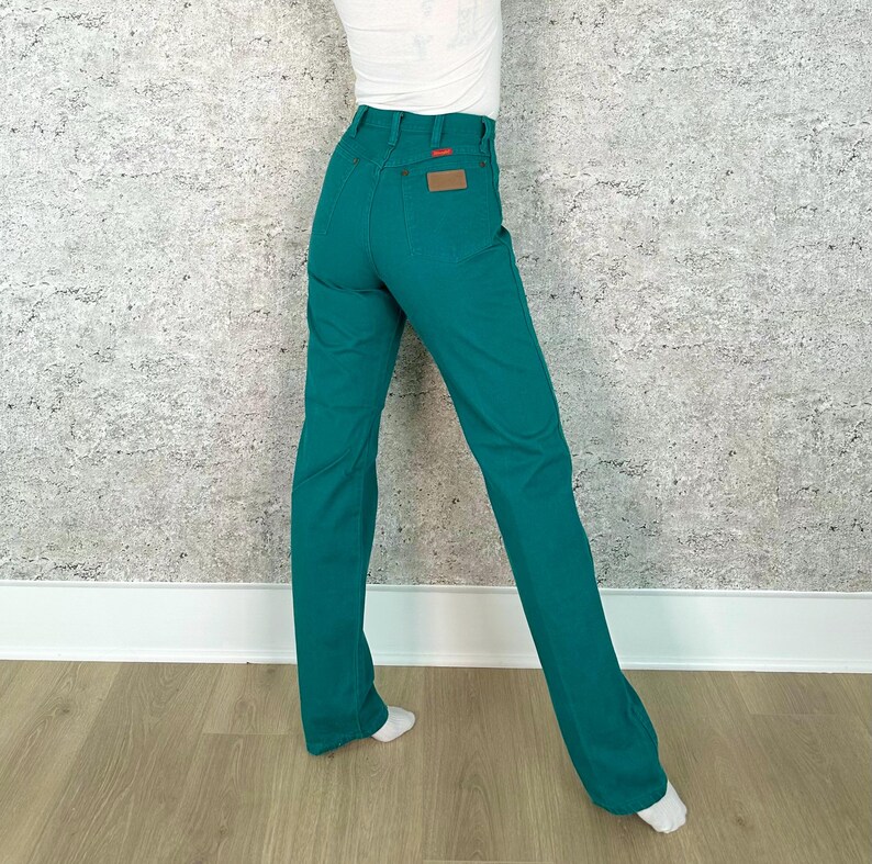 May include: Teal-colored denim pants with a high waist and a straight leg. The pants have a brown leather patch on the back pocket and a red tag. The pants are shown from the waist down.