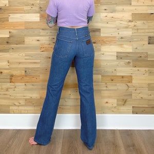 Pu&ograve; includere: Jeans Wrangler blu in denim con gamba svasata. I jeans hanno una toppa in pelle marrone sulla tasca posteriore e vita alta. La modella indossa un top corto lilla.