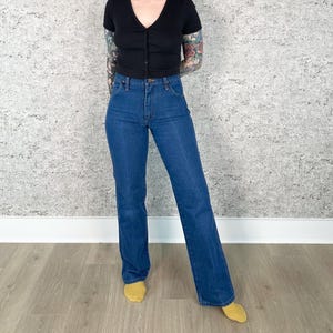 May include: A person wearing a black, short-sleeved, button-down top and blue denim jeans. The jeans have a classic, straight-leg fit. The person is wearing yellow socks.