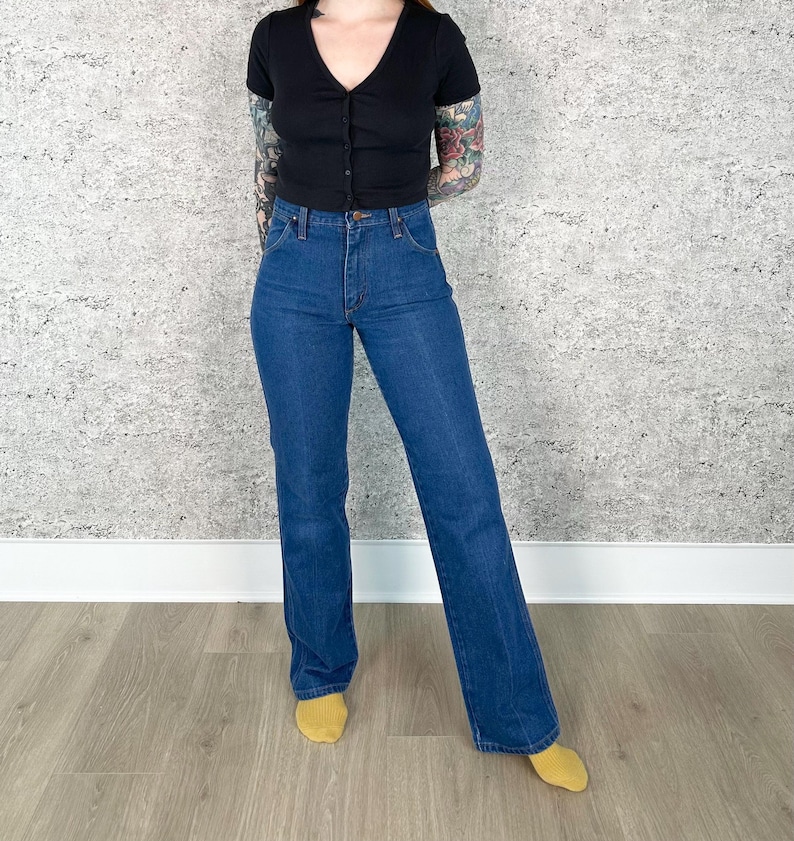 May include: A person wearing a black, short-sleeved, button-down top and blue denim jeans. The jeans have a classic, straight-leg fit. The person is wearing yellow socks.