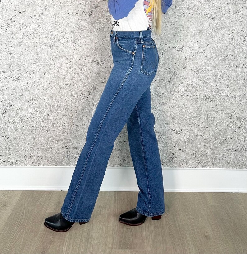 K&ouml;nnte beinhalten: Blaue Jeans aus Denim mit hoher Taille und ausgestelltem Bein. Die Jeans haben ein klassisches F&uuml;nf-Taschen-Design und werden mit schwarzen Lederstiefeln getragen. Die Jeans haben eine mittlere Waschung und scheinen Vintage zu sein.