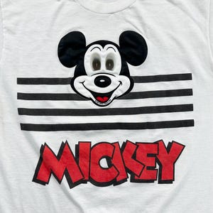 May include: White t-shirt featuring a smiling Mickey Mouse face above a black and white striped design. The word "MICKEY" is printed in red, outlined in black, across the bottom of the shirt.