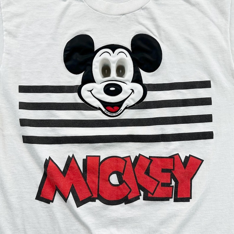 May include: White t-shirt featuring a smiling Mickey Mouse face above a black and white striped design. The word "MICKEY" is printed in red, outlined in black, across the bottom of the shirt.