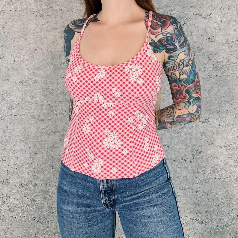 May include: A pink and white gingham camisole top with white daisy floral accents. The top has thin straps and a scoop neckline. The model is wearing blue jeans.