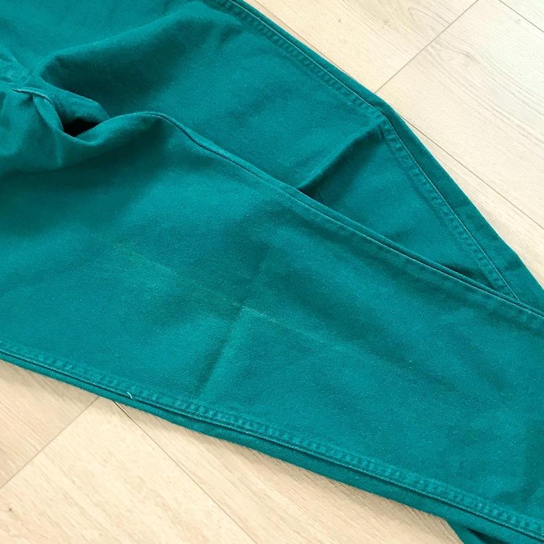 May include: Close-up of teal-colored denim pants. The pants are folded, showing the front pockets and seams. The fabric has a slight sheen, and the stitching is visible. The overall look is casual and comfortable.