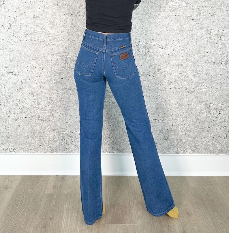 May include: Blue denim Wrangler jeans with a high waist and flared leg. The jeans have classic five-pocket styling, a brown leather patch on the back, and a zip fly. The jeans are a medium wash and appear to be vintage.