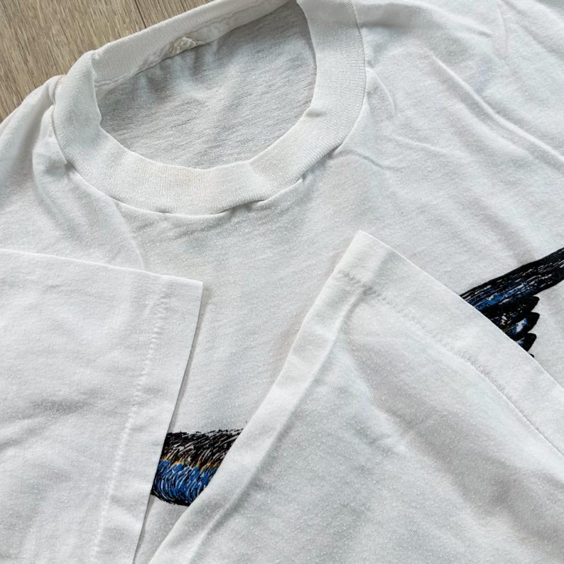 May include: White t-shirt with a crew neck and short sleeves. The shirt features a graphic of a bird with black, blue, and brown feathers on the right side. The shirt is made of a soft, likely cotton, material.