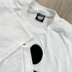 May include: White XL t-shirt with a black graphic design. The shirt has a crew neck and short sleeves. The tag reads "SCREEN STARS BEST" and "MADE IN USA". The shirt is made of 50% cotton and 50% polyester.