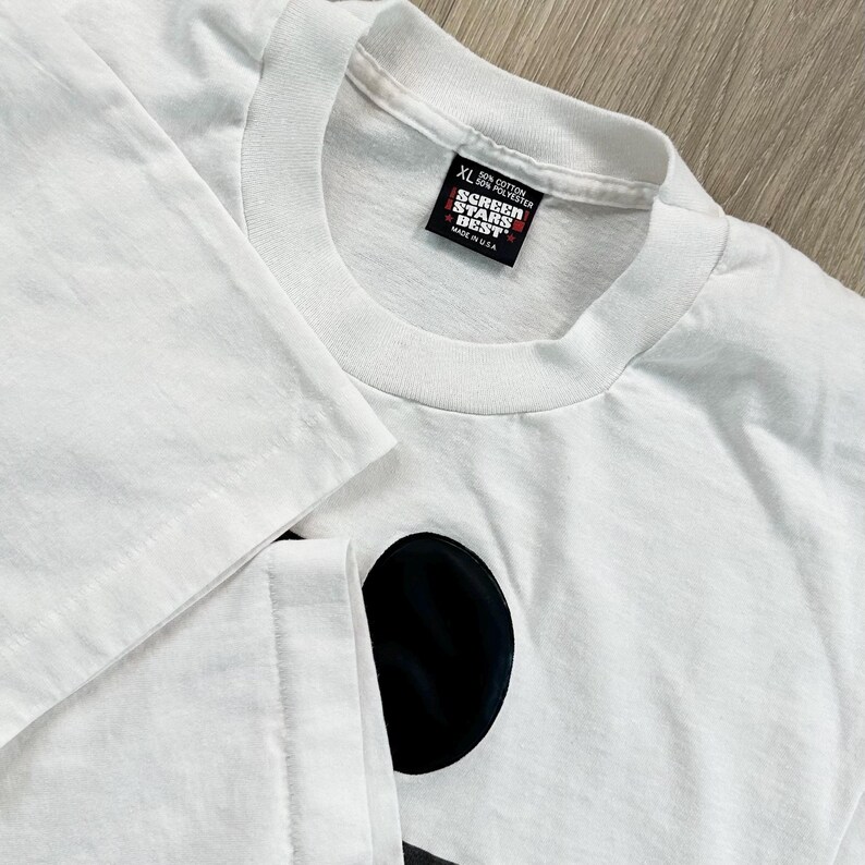 May include: White XL t-shirt with a black graphic design. The shirt has a crew neck and short sleeves. The tag reads "SCREEN STARS BEST" and "MADE IN USA". The shirt is made of 50% cotton and 50% polyester.