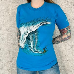 May include: A bright blue short-sleeve t-shirt featuring a large graphic print of a crashing ocean wave with white sea foam and a small surfer on a yellow surfboard at the base. The shirt has a crew neckline and a small logo on the sleeve.