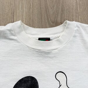 Op de afbeelding: Wit t-shirt met een zwart-wit cartoon karakter ontwerp. Het shirt heeft een ronde hals en korte mouwen. Het ontwerp is zichtbaar op het onderste deel van het shirt.
