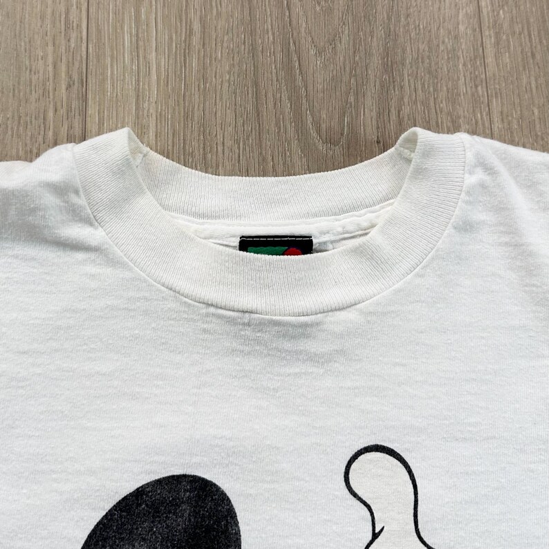 Op de afbeelding: Wit t-shirt met een zwart-wit cartoon karakter ontwerp. Het shirt heeft een ronde hals en korte mouwen. Het ontwerp is zichtbaar op het onderste deel van het shirt.