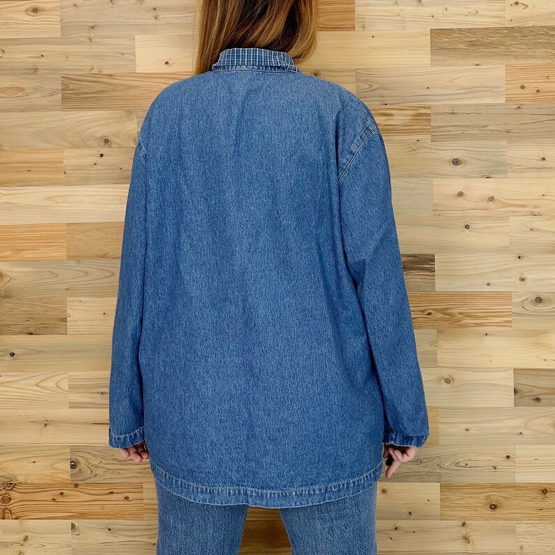 May include: A medium-wash denim jacket and matching jeans. The jacket has long sleeves and a collared neckline with a blue and white plaid pattern. The outfit is displayed against a light brown wood background.