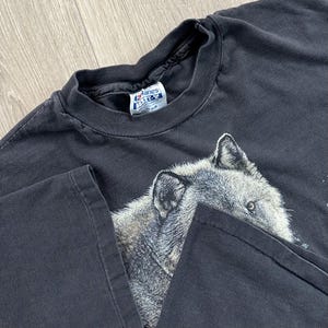 Peut inclure: T-shirt noir avec un motif d&eacute;taill&eacute; d'une t&ecirc;te de loup. Le loup est repr&eacute;sent&eacute; en nuances de gris et de blanc, avec des yeux per&ccedil;ants. Le t-shirt a un col rond classique et des manches courtes.