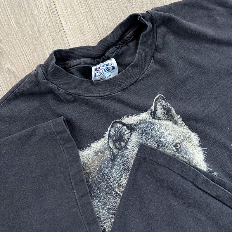 Peut inclure: T-shirt noir avec un motif d&eacute;taill&eacute; d'une t&ecirc;te de loup. Le loup est repr&eacute;sent&eacute; en nuances de gris et de blanc, avec des yeux per&ccedil;ants. Le t-shirt a un col rond classique et des manches courtes.