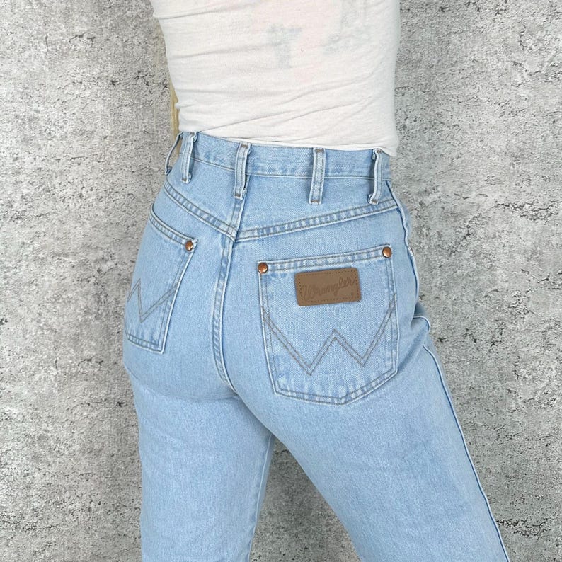 May include: Pale blue denim jeans with a classic fit. Features include a brown leather patch with the "Wrangler" logo on the back pocket, copper rivets, and a high waist. The jeans have a traditional five-pocket design and a straight leg.