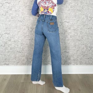 K&ouml;nnte beinhalten: Hellblaue Wrangler Jeans aus Denim mit hoher Taille und geradem Bein. Die Jeans hat einen braunen Lederaufn&auml;her auf der Ges&auml;&szlig;tasche. Die Person tr&auml;gt ein wei&szlig;es und blaues Langarmshirt mit dem Text "Bite the Dust Tour".