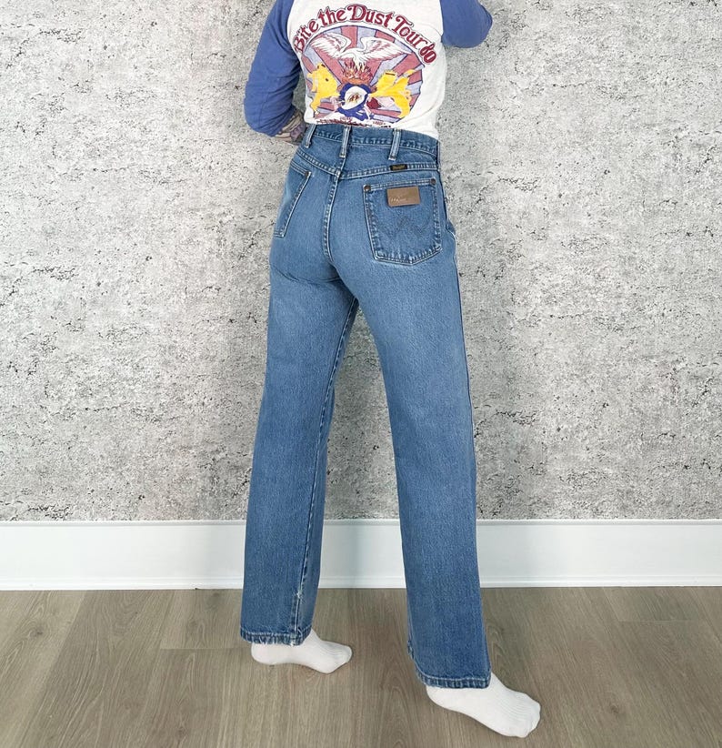 K&ouml;nnte beinhalten: Hellblaue Wrangler Jeans aus Denim mit hoher Taille und geradem Bein. Die Jeans hat einen braunen Lederaufn&auml;her auf der Ges&auml;&szlig;tasche. Die Person tr&auml;gt ein wei&szlig;es und blaues Langarmshirt mit dem Text "Bite the Dust Tour".