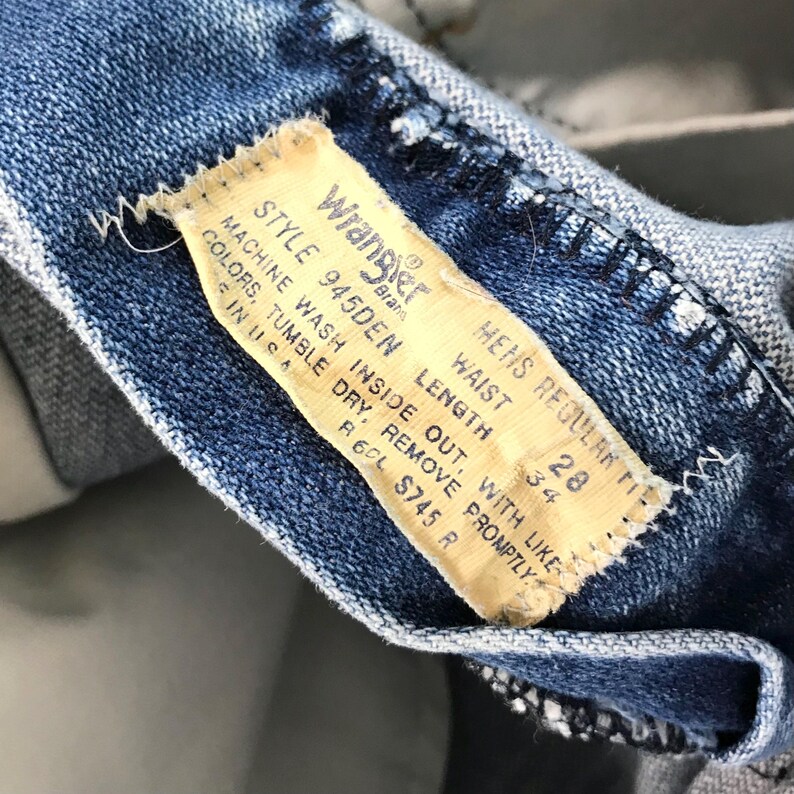 Op de afbeelding: Close-up van een blauwe denim jeans met een Wrangler merklabel. Het label geeft een taillemaat van 71 cm en een lengte van 86 cm aan. De jeans is stijl 945DEN.
