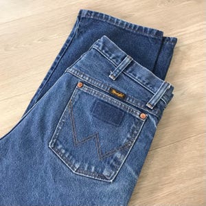 K&ouml;nnte beinhalten: Blaue Jeans aus Denim mit dem Wrangler-Logo auf der Ges&auml;&szlig;tasche. Die Jeans ist gefaltet und zeigt die Ges&auml;&szlig;taschen und G&uuml;rtelschlaufen. Die N&auml;hte sind sichtbar, und die Jeans scheint in gutem Zustand zu sein.