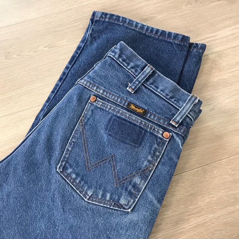 K&ouml;nnte beinhalten: Blaue Jeans aus Denim mit dem Wrangler-Logo auf der Ges&auml;&szlig;tasche. Die Jeans ist gefaltet und zeigt die Ges&auml;&szlig;taschen und G&uuml;rtelschlaufen. Die N&auml;hte sind sichtbar, und die Jeans scheint in gutem Zustand zu sein.