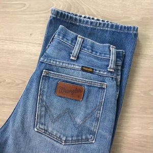 Op de afbeelding: Lichtblauwe spijkerbroek met het Wrangler-logo op de achterzak. De jeans heeft een klassiek ontwerp met vijf zakken, riemlussen en een knoop- en ritssluiting. De jeans is opgevouwen en lijkt in goede staat te verkeren.