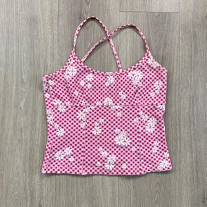 May include: A pink gingham camisole top with white daisy floral print. The top features a criss-cross back design and thin straps. The fabric has a checkered pattern.