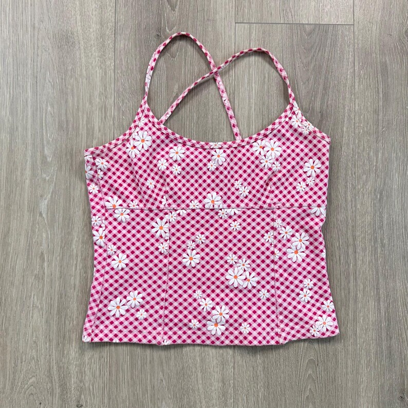 May include: A pink gingham camisole top with white daisy floral print. The top features a criss-cross back design and thin straps. The fabric has a checkered pattern.