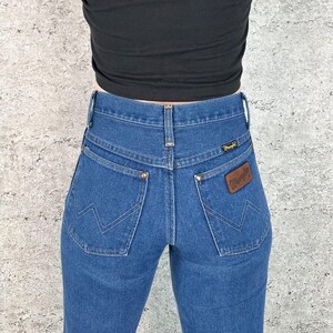 May include: Blue denim jeans with a high waist and a classic five-pocket design. The jeans feature the Wrangler logo on a brown leather patch and on the back pocket. The jeans are a medium wash and have a straight leg.