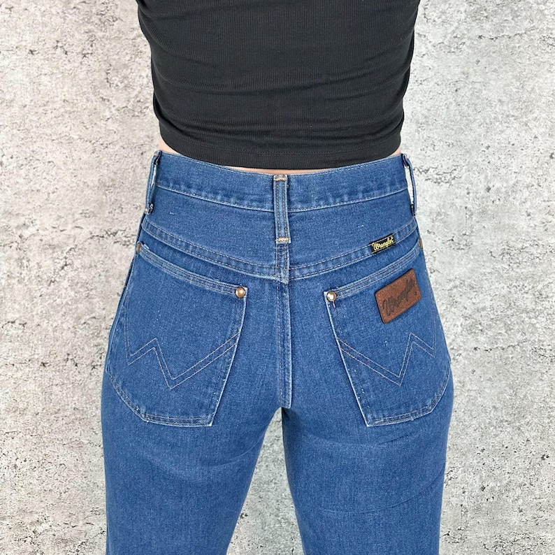 May include: Blue denim jeans with a high waist and a classic five-pocket design. The jeans feature the Wrangler logo on a brown leather patch and on the back pocket. The jeans are a medium wash and have a straight leg.
