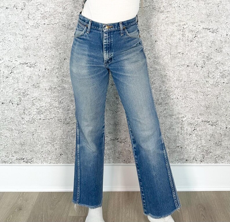 May include: Blue denim jeans with a button and zip fastening. The jeans have a classic five-pocket design and a straight leg fit. The hem is frayed. The jeans are worn by a person.