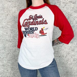 Peut inclure: T-shirt de baseball blanc et rouge de style raglan. Le t-shirt porte l'inscription "St. Louis Cardinals WORLD SERIES 1985" avec un graphisme de cardinal. Les manches sont rouges et le corps est blanc.