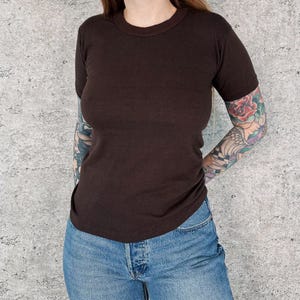 May include: A solid dark brown short-sleeve t-shirt with a crew neckline, paired with blue denim jeans. The t-shirt features a simple, plain design with a fitted silhouette, constructed from a smooth fabric, and worn with classic blue denim pants.