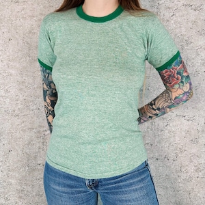 Puede incluir: Una camiseta verde claro con cuello y ribetes de manga verdes. La camiseta está hecha de un material suave y texturizado y se combina con unos vaqueros azules. El estilo general es casual y vintage.