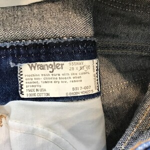 May include: A blue denim Wrangler jeans label with white text. The label says "935NAV 28 x 33 30", "Machine wash warm with like colours, only non-chlorine bleach when needed, tumble dry low, remove promptly", "MADE IN USA", "B317-007", "0 84084 404275", and "100% COTTON".