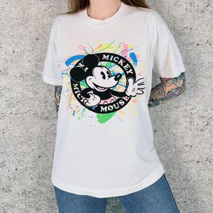 Może przedstawiać: Biała koszulka z grafiką Myszki Miki w czarnym okręgu z napisem "Mickey Mouse". Projekt zawiera kolorowe plamy farby. Koszulka ma krótkie rękawy i okrągły dekolt.