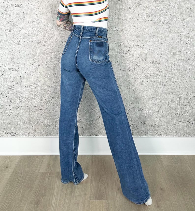 Puede incluir: Vaqueros de mezclilla azules con cintura alta y perneras acampanadas. Los vaqueros tienen un dise&ntilde;o cl&aacute;sico de cinco bolsillos y un lavado oscuro. La modelo lleva una camiseta a rayas. Los vaqueros son de estilo vintage.