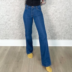 May include: Dark blue denim jeans with a high waist and flared leg. The jeans have a classic five-pocket design and a button and zipper closure. The jeans are a medium wash and appear to be vintage.