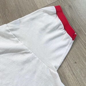May include: Close-up of a white t-shirt sleeve with a red trim around the cuff. The fabric appears soft and slightly textured. The sleeve is angled, showing the seam and the contrast between the white body and the red cuff.