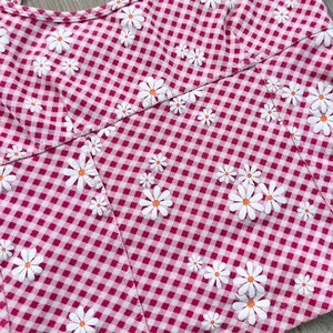 May include: Close-up of a pink and white gingham fabric with white daisy flower appliqu&eacute;s. The daisies have orange centres. The fabric is cut in a bodice shape, suggesting a garment.