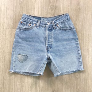 May include: Light blue denim shorts with a frayed hem. The shorts feature a heart-shaped patch on the left leg. Classic five-pocket design with a button and zipper closure. A casual, everyday wear item.