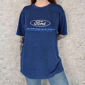 May include: A navy blue t-shirt with the Ford logo in white and the word "MOTORSPORT" in blue. The shirt has short sleeves and a crew neck. The fabric appears to be a soft, comfortable material.