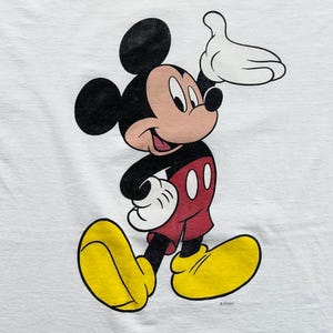 Op de afbeelding: Een wit t-shirt met een cartoonafbeelding van Mickey Mouse. Mickey wordt afgebeeld terwijl hij loopt, met zijn rechterhand omhoog. Hij draagt een rode broek, gele schoenen en witte handschoenen. Het is een vintage ontwerp.