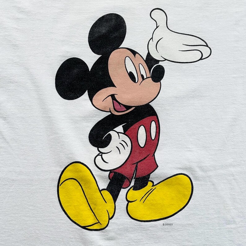 Op de afbeelding: Een wit t-shirt met een cartoonafbeelding van Mickey Mouse. Mickey wordt afgebeeld terwijl hij loopt, met zijn rechterhand omhoog. Hij draagt een rode broek, gele schoenen en witte handschoenen. Het is een vintage ontwerp.