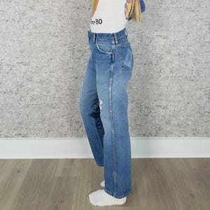 K&ouml;nnte beinhalten: Hellblaue Jeans mit geradem Bein. Die Jeans haben ein klassisches F&uuml;nf-Taschen-Design, einen Knopf- und Rei&szlig;verschluss sowie Used-Details. Die Jeans werden mit einem wei&szlig;en T-Shirt mit der Aufschrift "80" getragen.